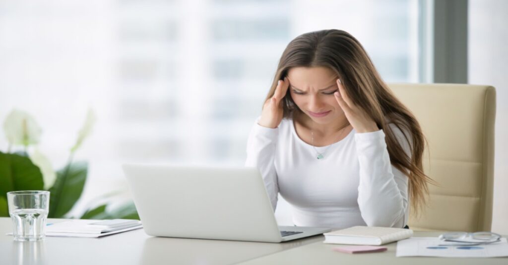 Female sales rep in front of laptop with head in hands, frustrated by ERP legacy system issues.